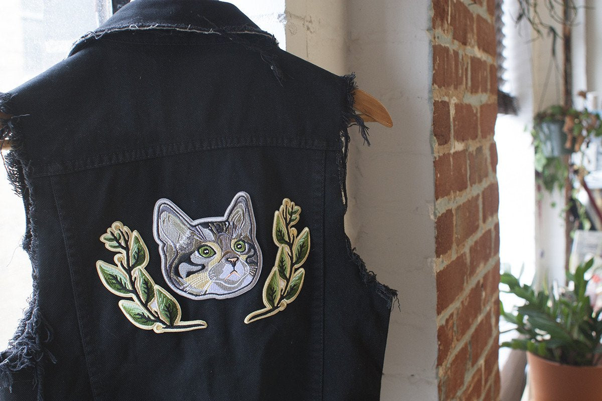 A pair of green laurel leaf patches and a large cat face patch, attached to a black denim waistcoat
