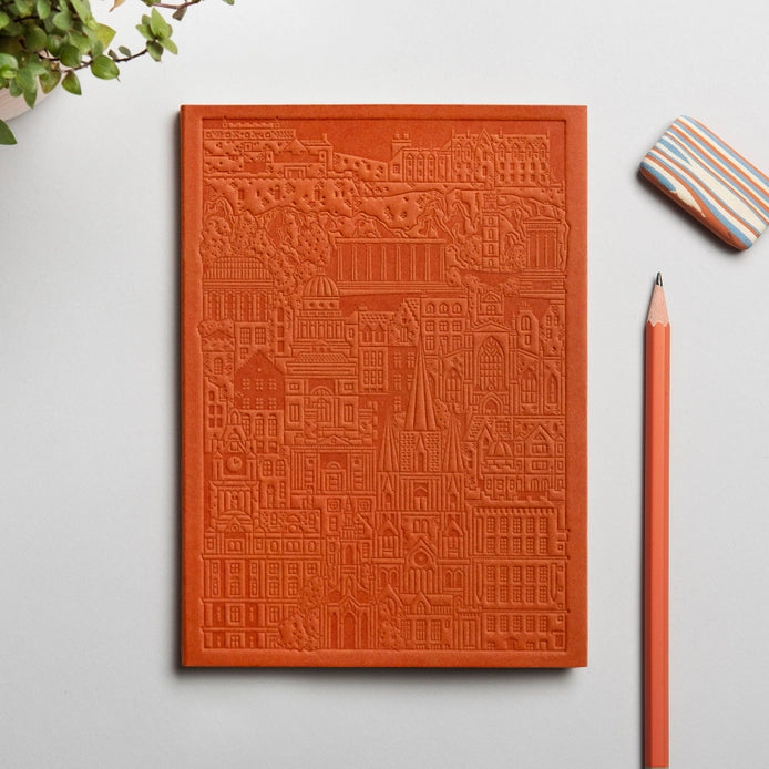 A hand-pressed orange notebook with a cityscape embossed on the cover, accompanied by a pencil and eraser, displayed on a white background.