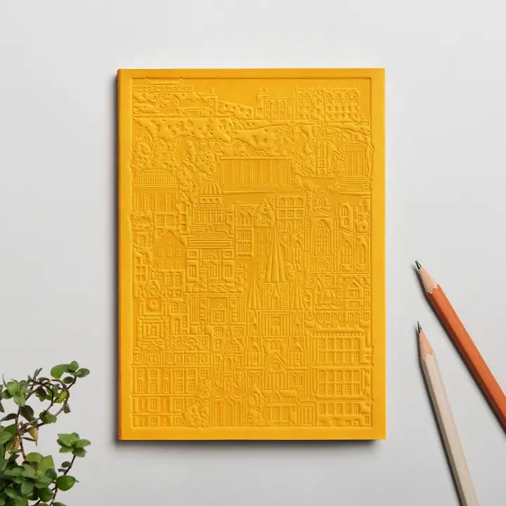 A yellow notebook with a hand-pressed embossed cover of Edinburgh buildings, displayed alongside a plant and pencils on a white background.