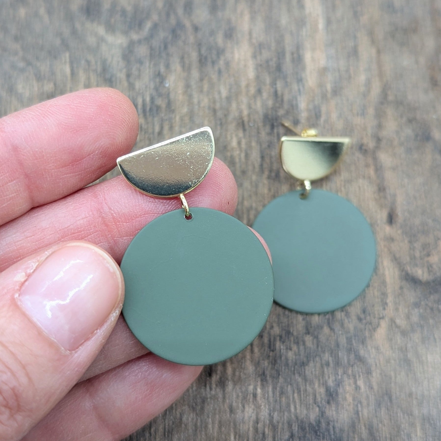 Green earrings with gold accents held by a hand on a wooden surface