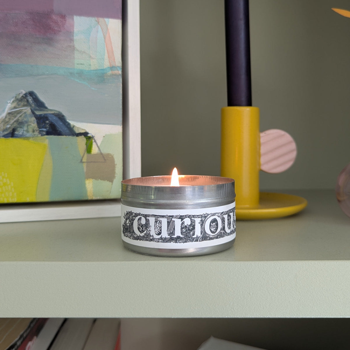 A lit silver tin candle with a black and white patterned label reading 'curious' sits on a light sage green shelf, styled with a yellow and navy candlestick holder and framed art.