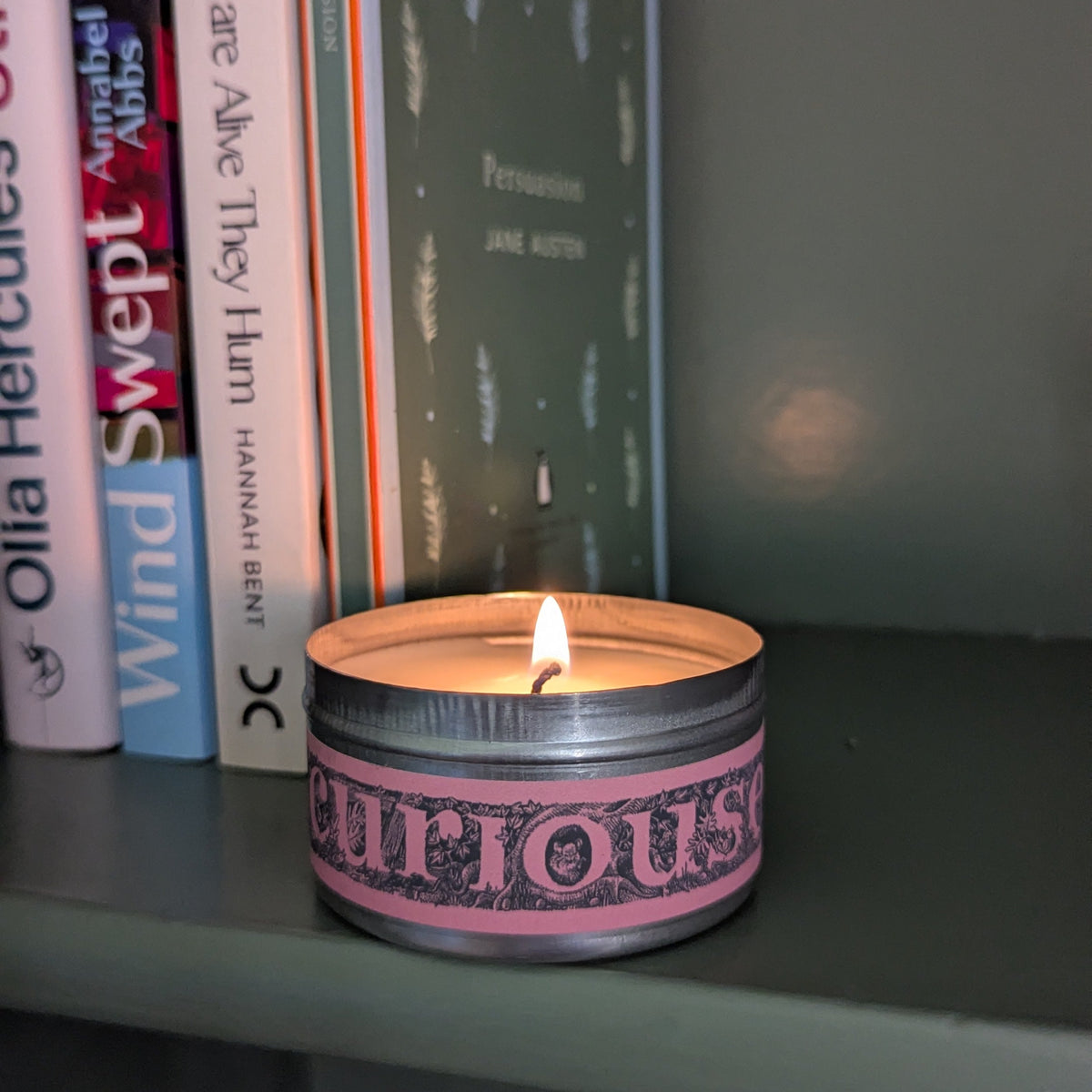 A lit tin candle with a pink and black patterned label reading 'CURIIOUS' sits on a shelf next to several books.