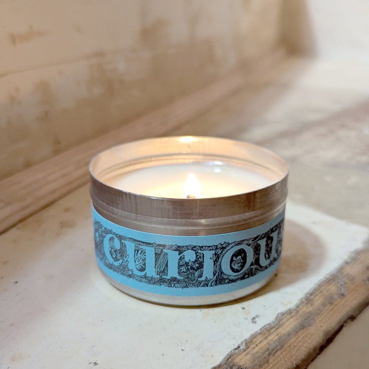A lit tealight candle in a small round tin with a light blue and black patterned label reading 'curious', placed on a worn wooden surface.