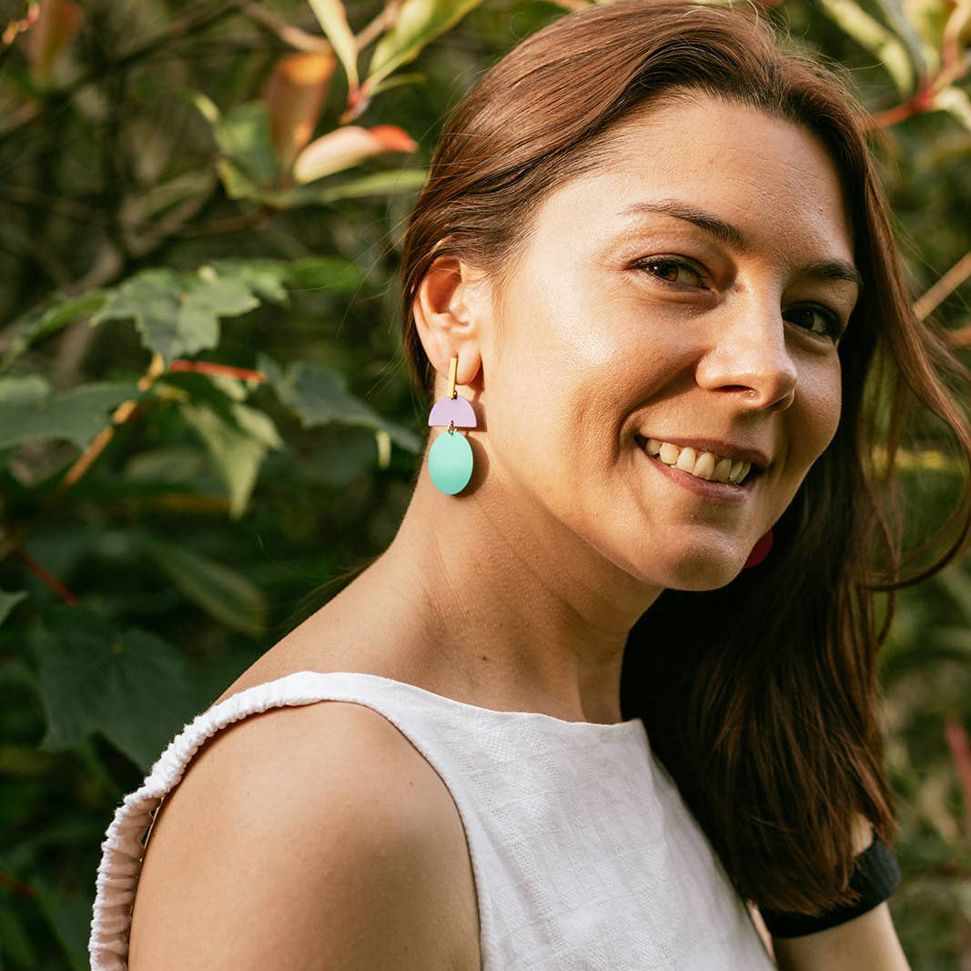 A woman with brown hair wearing a white sleeveless top and a pastel pink and mint green geometric drop earring, posing outdoors in front of green foliage.