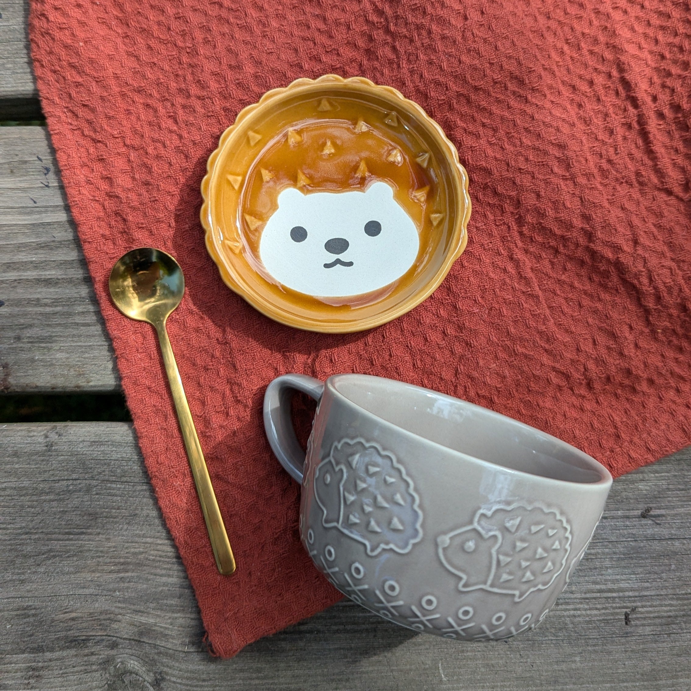 Ceramic cup with a hedgehog design, spoon, and textured mug on a red cloth over a wooden surface