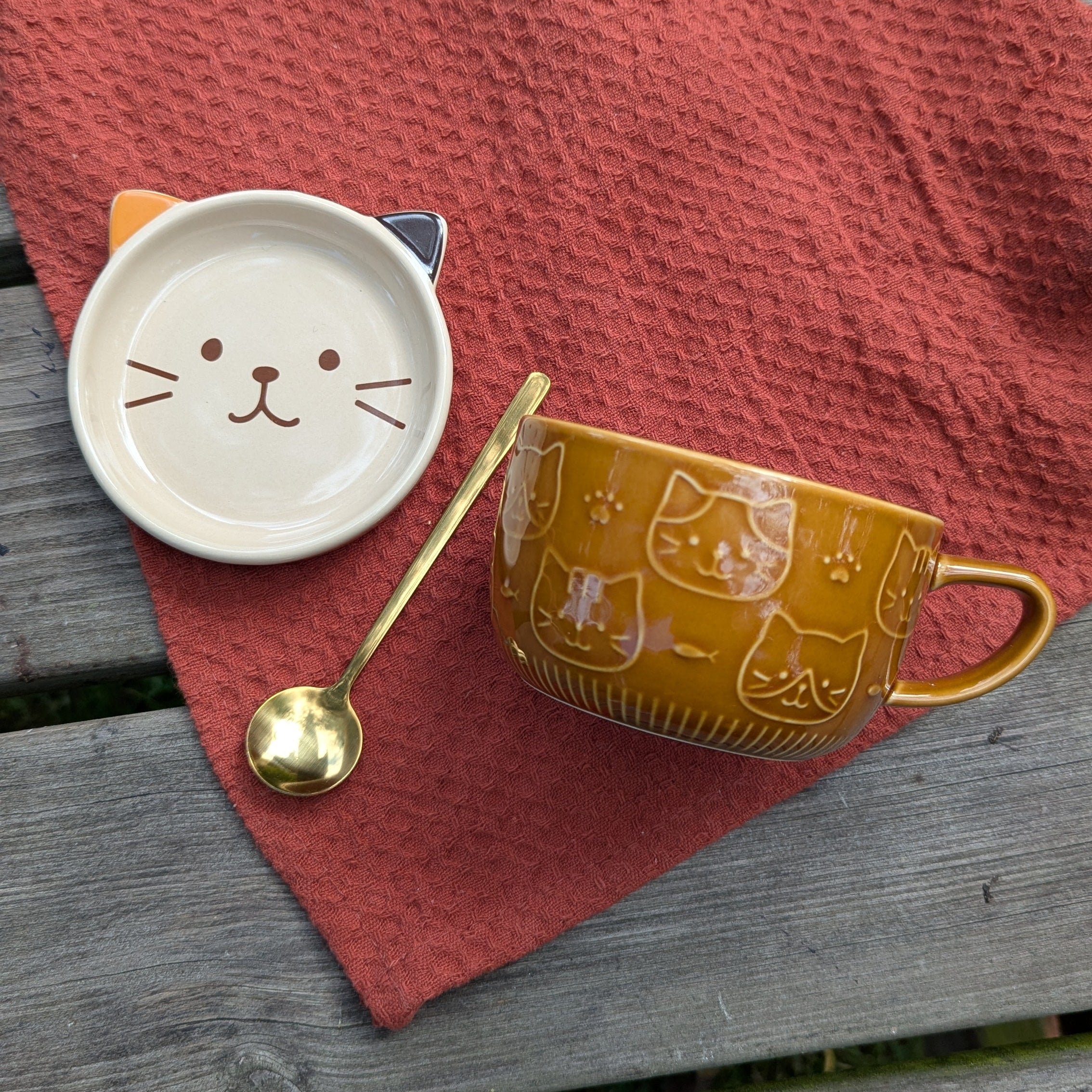 Cat-themed ceramic cup, saucer, and spoon on a red cloth over a wooden surface