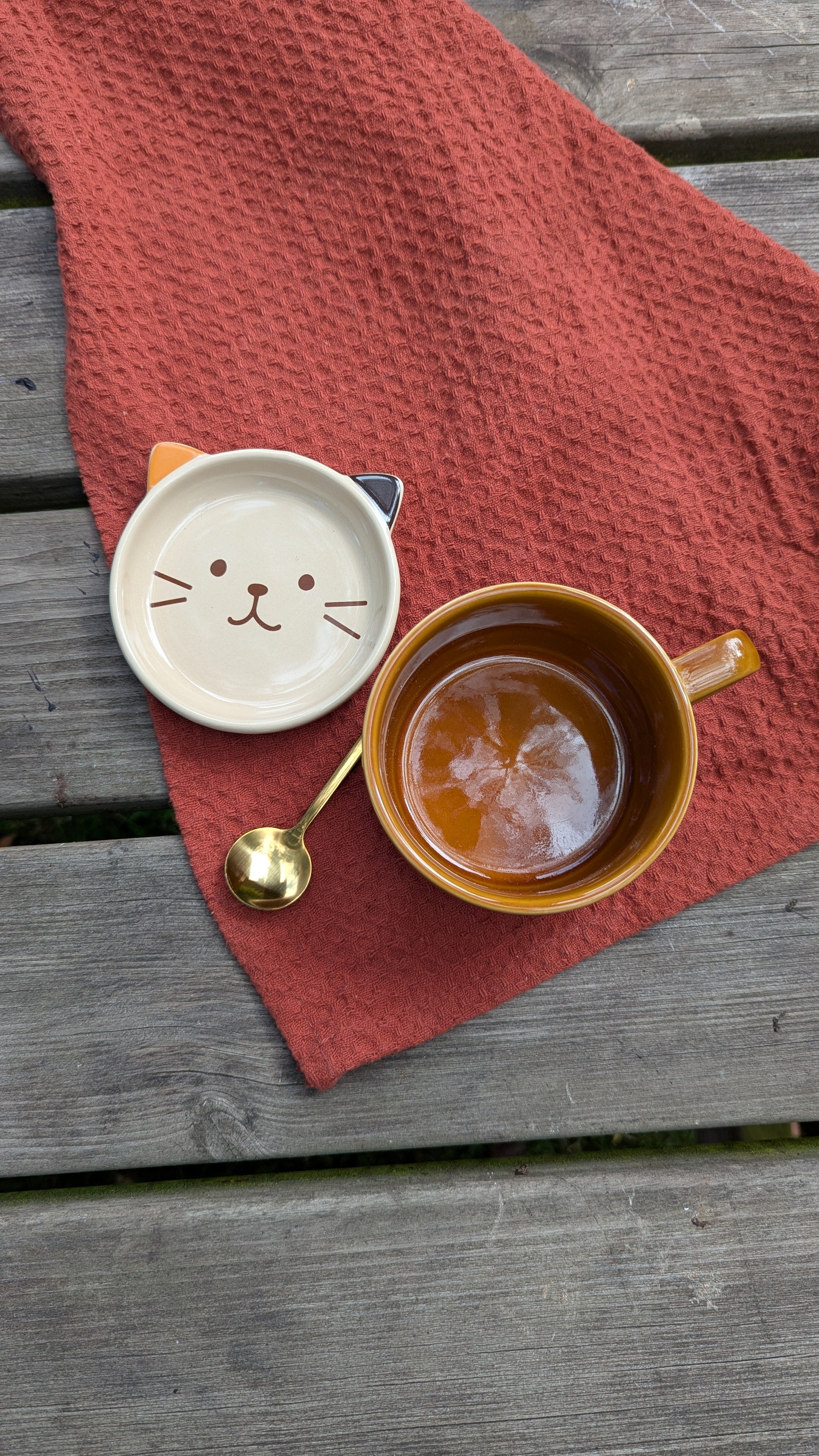 Ceramic mug with saucer with cat face, spoon on a red cloth over a wooden surface