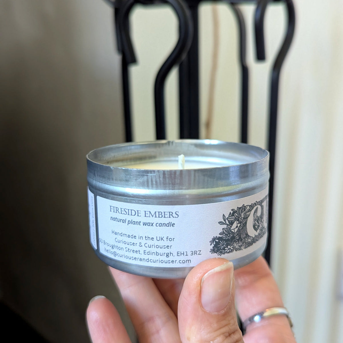 A hand holding a small silver tin candle labeled 'FIRESIDE EMBERS' against a softly blurred indoor background with a black metal stand.