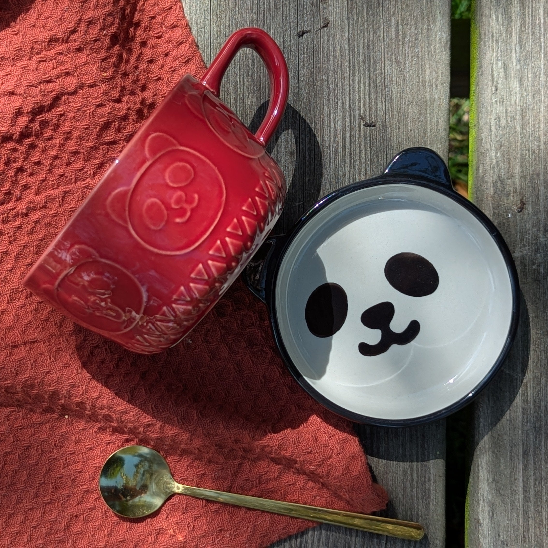 Red mug with a panda design, black and white saucer that is a lid with a smiley face, and gold spoon on a red fabric background.