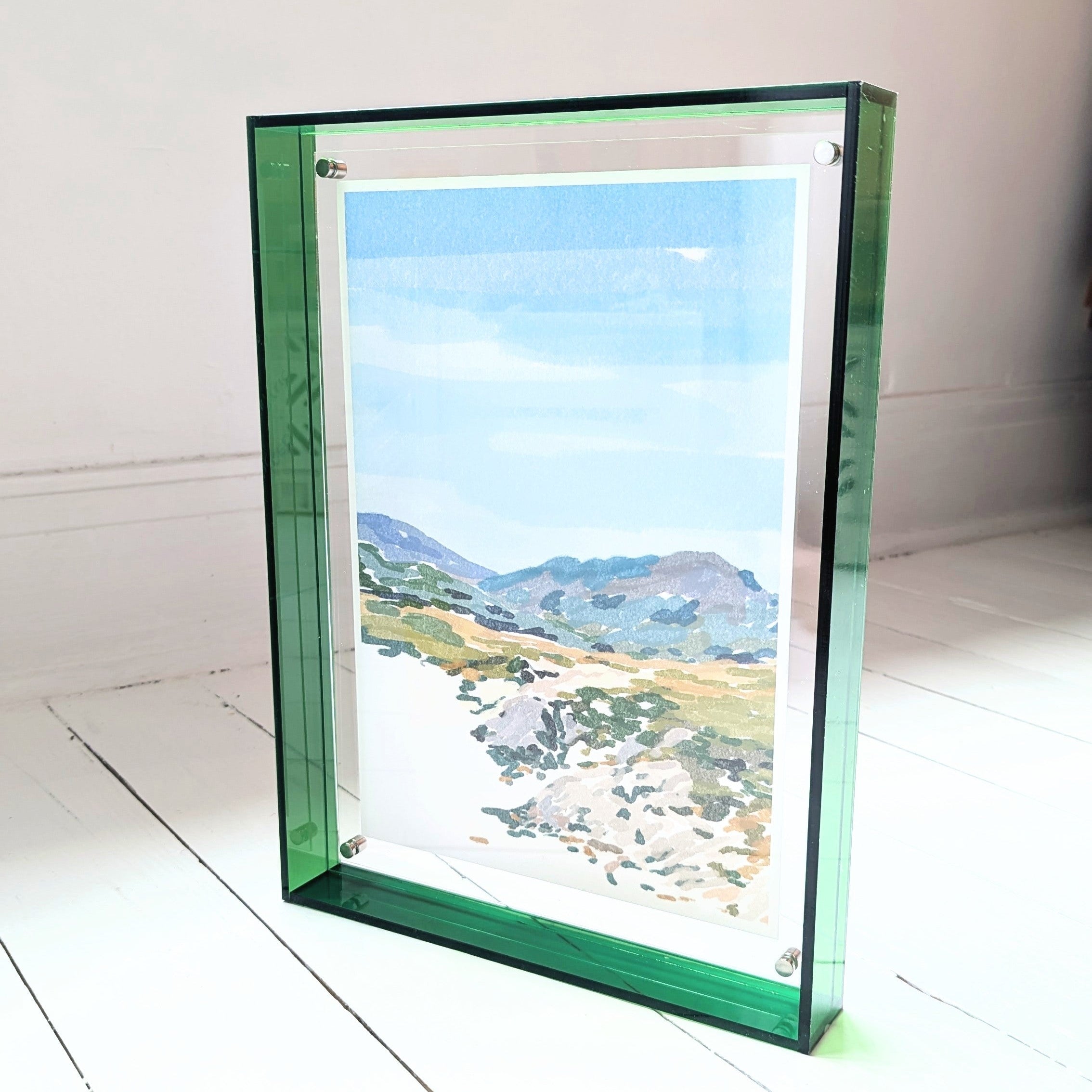 Framed artwork of a landscape with mountains and a beach on a white surface.