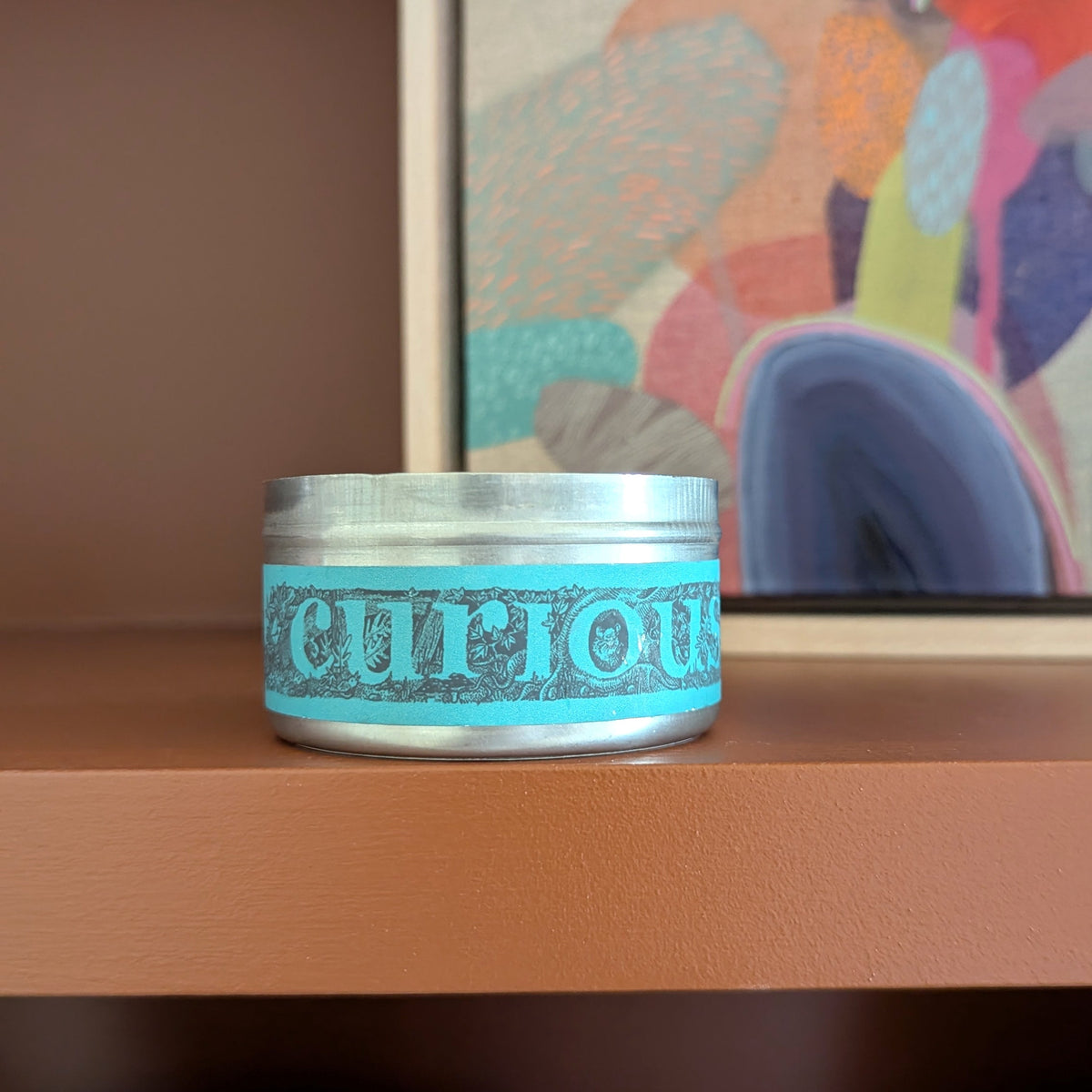 A small round metal tin candle with a turquoise blue label reading 'curious' sits on a wooden shelf in front of a colorful abstract painting.