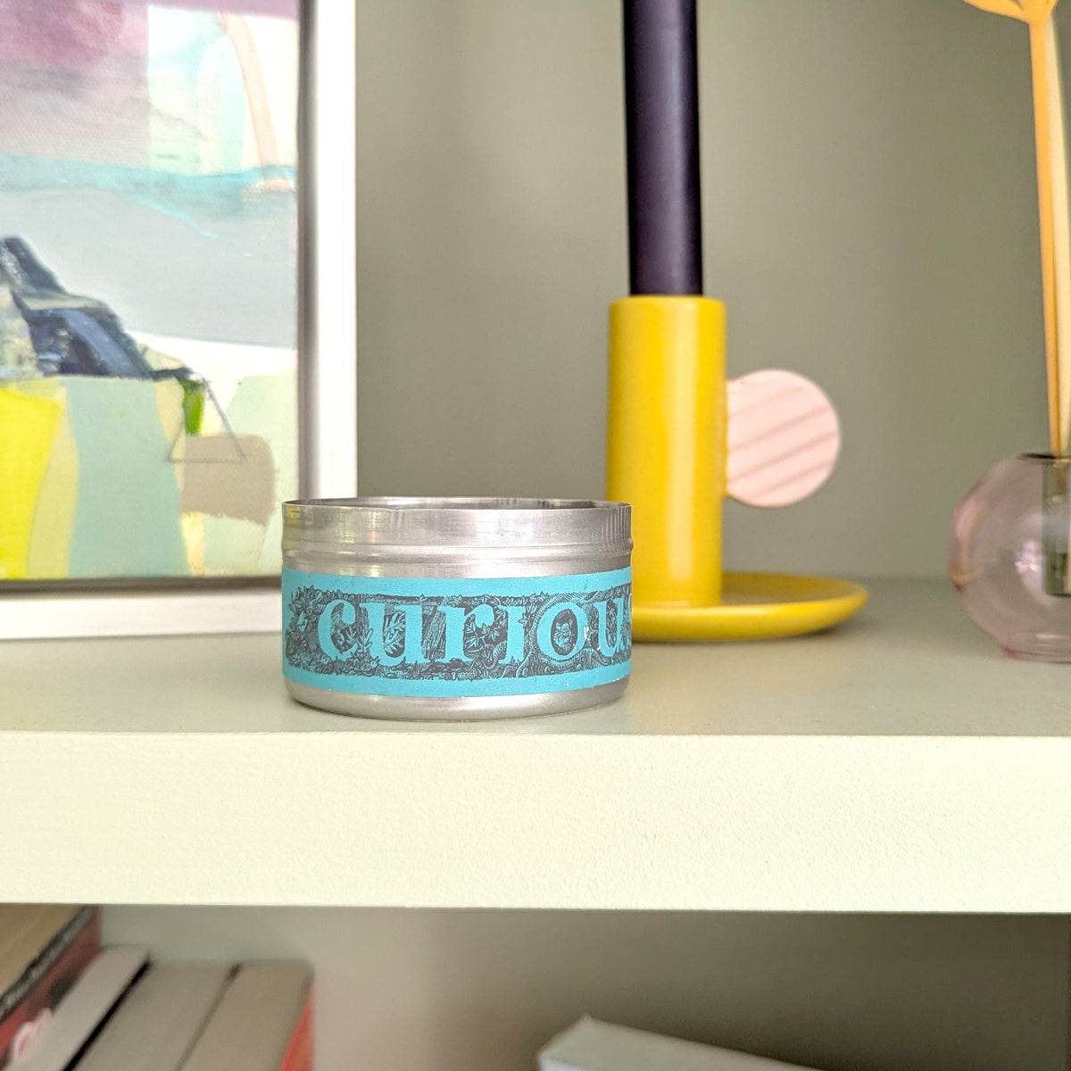 A round tin candle with a turquoise blue label reading 'curious' sits on a light shelf next to a yellow and navy candle holder, with framed art and books in the background.