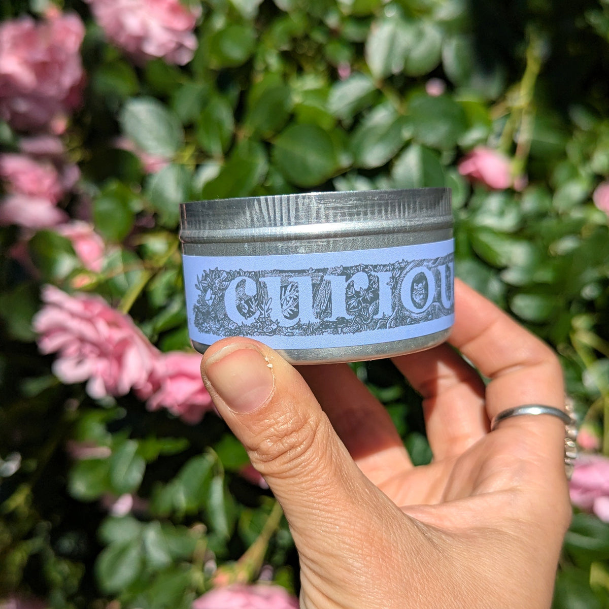 A hand holding a small jar labeled 'curious' in front of a background of green leaves and pink flowers.