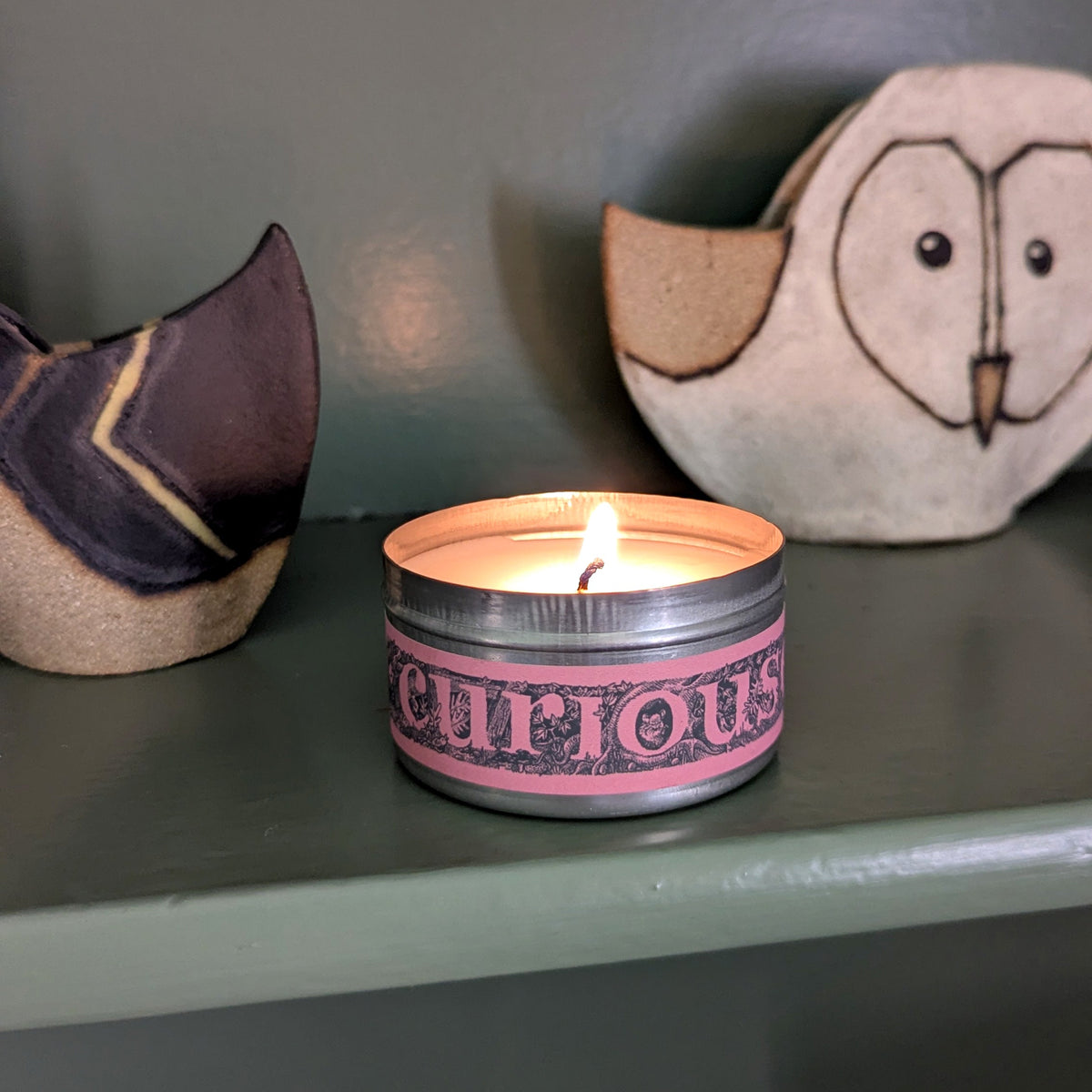 A lit silver tin candle with a pink label reading 'CURIOUS' sits on a dark green shelf, flanked by decorative dried flowers against a dark green wall.